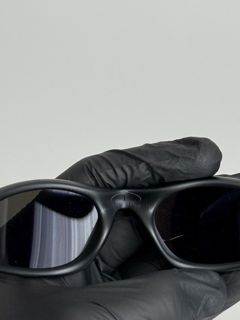 May include: Black sunglasses with a rounded frame and dark lenses. The sunglasses have the word "MINUTE" printed on the top of the frame. The sunglasses are held by a person wearing black gloves.
