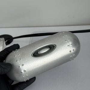 May include: Silver-colored eyewear case with a riveted design. The case has a rounded shape and features the Oakley logo in black. The case is held by a gloved hand.
