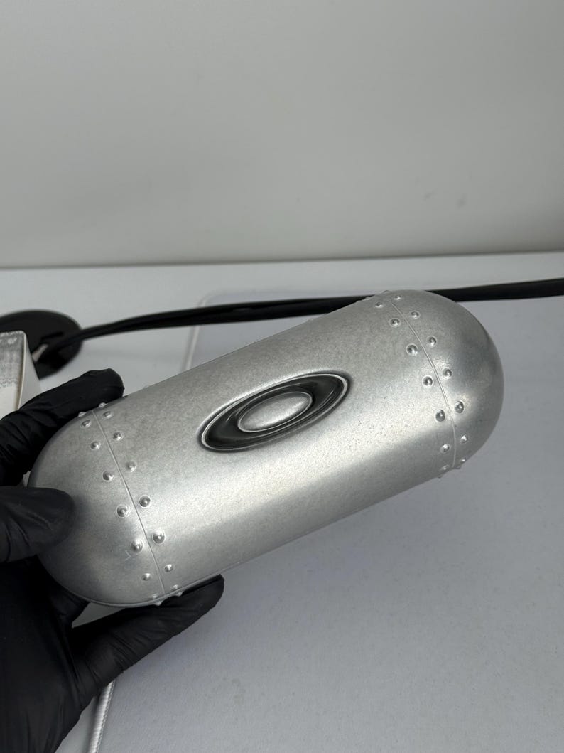 May include: Silver-colored eyewear case with a riveted design. The case has a rounded shape and features the Oakley logo in black. The case is held by a gloved hand.