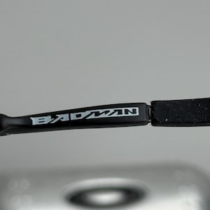 May include: Close-up of a black eyeglass frame with the word "BADMAN" in white lettering. The frame is sleek and modern, with a rectangular shape. The background is a blurred, neutral color.