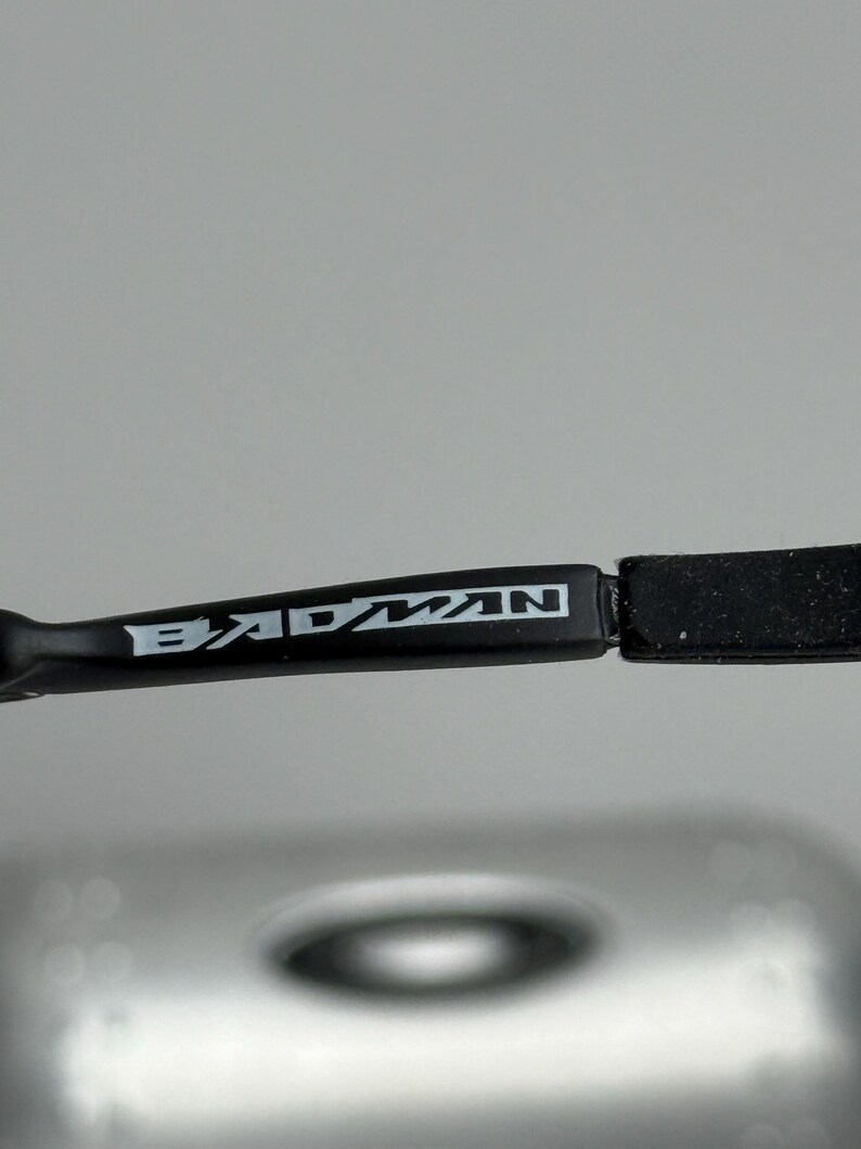 May include: Close-up of a black eyeglass frame with the word "BADMAN" in white lettering. The frame is sleek and modern, with a rectangular shape. The background is a blurred, neutral color.