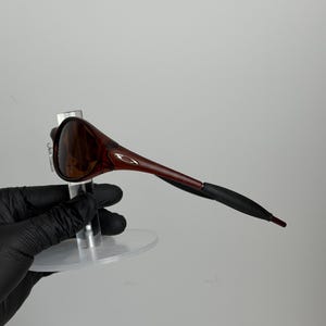 May include: Brown and black sunglasses with a sleek, modern design. The sunglasses have a dark brown frame and lenses, with black earpieces. The sunglasses are displayed on a clear stand.