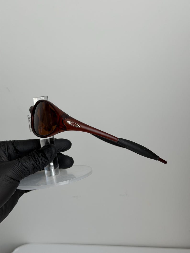 May include: Brown and black sunglasses with a sleek, modern design. The sunglasses have a dark brown frame and lenses, with black earpieces. The sunglasses are displayed on a clear stand.