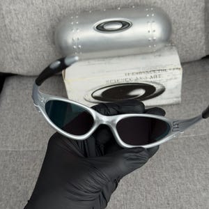 May include: Silver-framed sunglasses with dark lenses. The sunglasses are held in a black gloved hand. A silver case and a box with the text "SCIENCE AND ART" are in the background.