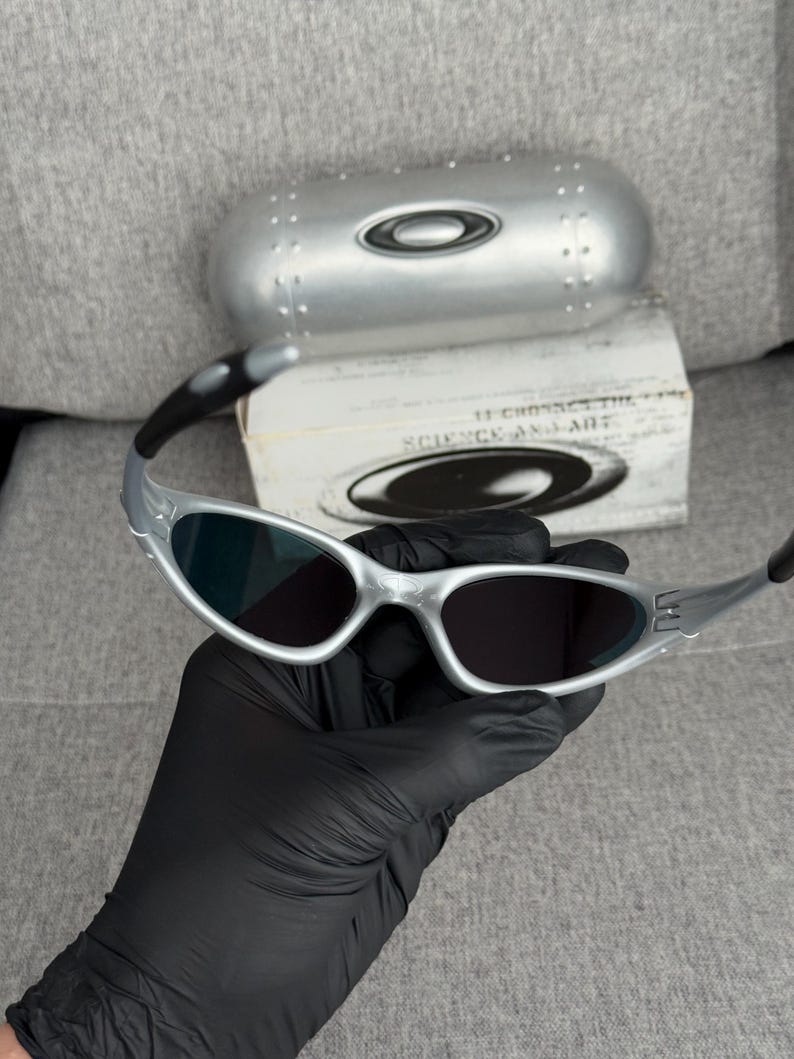 May include: Silver-framed sunglasses with dark lenses. The sunglasses are held in a black gloved hand. A silver case and a box with the text "SCIENCE AND ART" are in the background.
