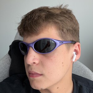 May include: Purple oval-shaped sunglasses with dark lenses. The sunglasses have a sleek, sporty design. The frame is a vibrant purple color. The lenses reflect the surroundings. The sunglasses are a stylish accessory.