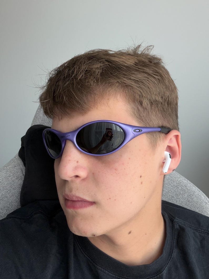 May include: Purple oval-shaped sunglasses with dark lenses. The sunglasses have a sleek, sporty design. The frame is a vibrant purple color. The lenses reflect the surroundings. The sunglasses are a stylish accessory.