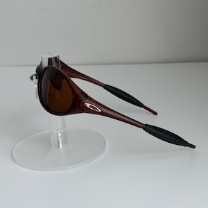 May include: Brown sunglasses with a dark frame and lenses. The sunglasses have a sleek design with a small silver logo on the side. The arms are a darker brown with black tips. The sunglasses are displayed on a clear stand.