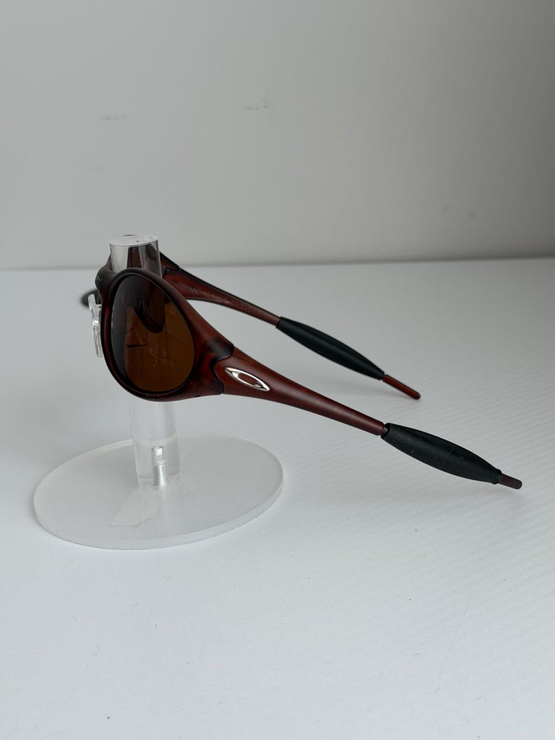 May include: Brown sunglasses with a dark frame and lenses. The sunglasses have a sleek design with a small silver logo on the side. The arms are a darker brown with black tips. The sunglasses are displayed on a clear stand.