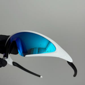 Puede incluir: Gafas de sol deportivas blancas y negras con lentes azules espejadas. Las gafas tienen un dise&ntilde;o elegante y aerodin&aacute;mico con montura blanca y patillas negras. Las lentes reflejan un tono azul brillante, para actividades al aire libre.
