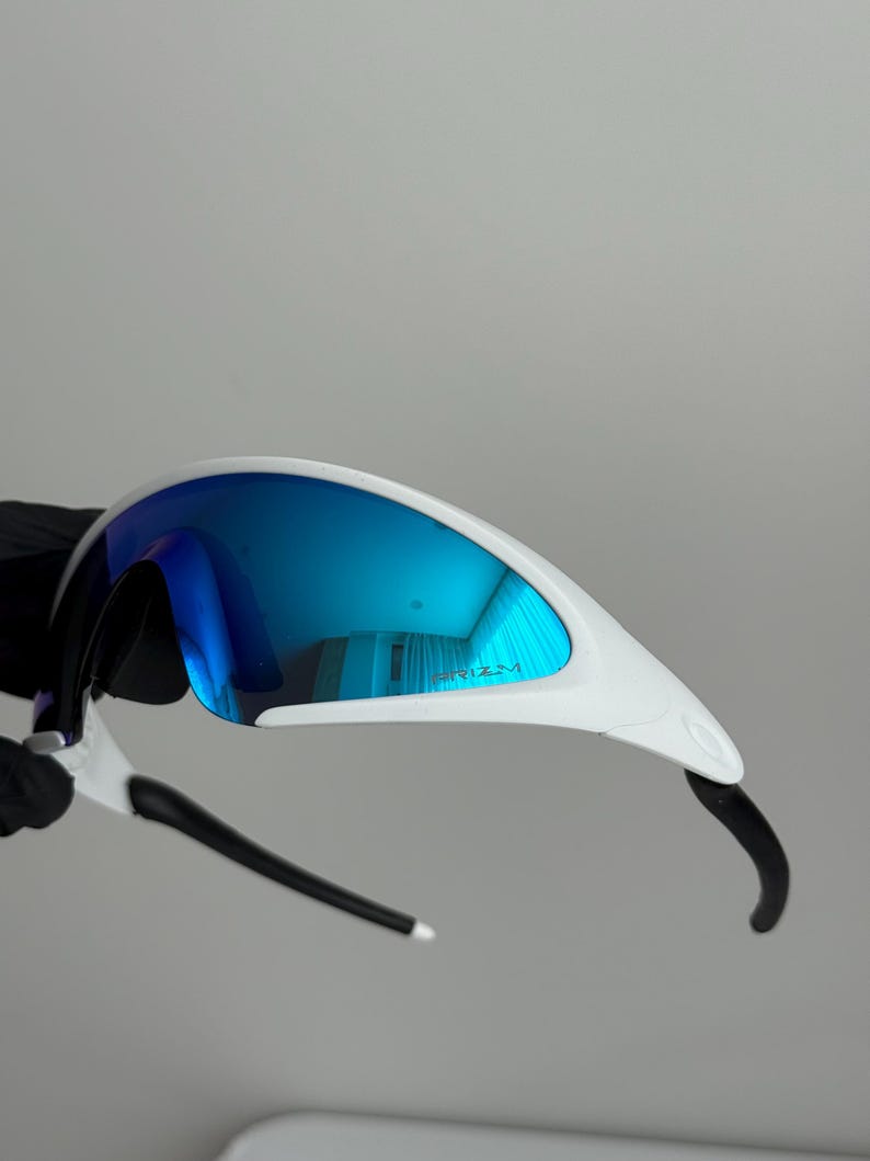 Puede incluir: Gafas de sol deportivas blancas y negras con lentes azules espejadas. Las gafas tienen un dise&ntilde;o elegante y aerodin&aacute;mico con montura blanca y patillas negras. Las lentes reflejan un tono azul brillante, para actividades al aire libre.