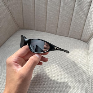 May include: Black sunglasses with a curved frame and dark lenses. The sunglasses have a small silver logo on the temple. The image shows the sunglasses held in a hand.