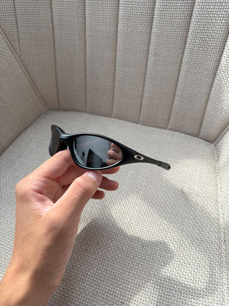 May include: Black sunglasses with a curved frame and dark lenses. The sunglasses have a small silver logo on the temple. The image shows the sunglasses held in a hand.