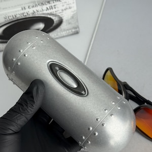 May include: A silver-colored, cylindrical sunglasses case with the Oakley logo. The case has a riveted design and is held by a gloved hand. A pair of sunglasses with orange lenses is visible.