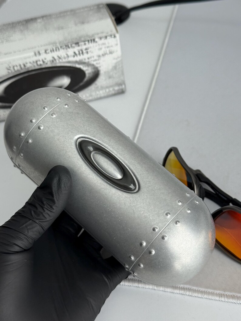 May include: A silver-colored, cylindrical sunglasses case with the Oakley logo. The case has a riveted design and is held by a gloved hand. A pair of sunglasses with orange lenses is visible.