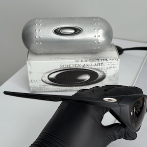 Puede incluir: Gafas de sol negras con acabado mate, sujetas por una mano enguantada. Las gafas tienen un dise&ntilde;o elegante y moderno. Un estuche plateado y una caja con el texto "SCIENCE AND ART" est&aacute;n en el fondo.