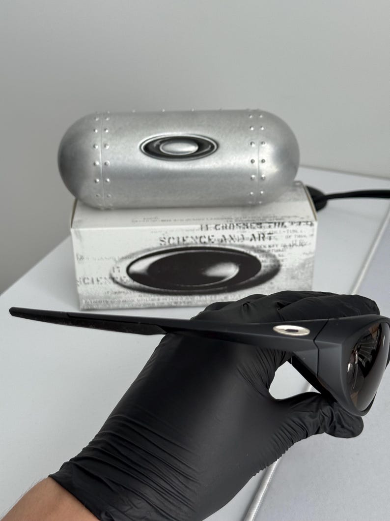 Puede incluir: Gafas de sol negras con acabado mate, sujetas por una mano enguantada. Las gafas tienen un dise&ntilde;o elegante y moderno. Un estuche plateado y una caja con el texto "SCIENCE AND ART" est&aacute;n en el fondo.