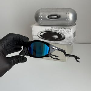 May include: Black sunglasses with blue mirrored lenses. The sunglasses have a unique, angular design. A silver case and a white box with the text "SCIENCE AND ART" are in the background.