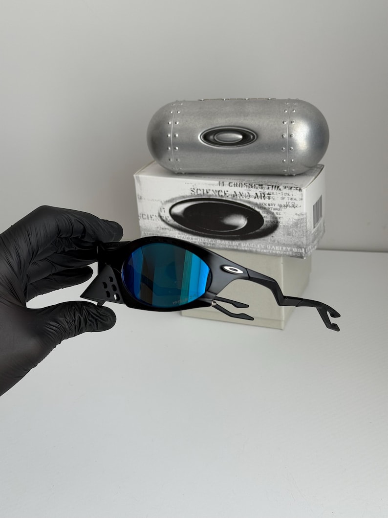 May include: Black sunglasses with blue mirrored lenses. The sunglasses have a unique, angular design. A silver case and a white box with the text "SCIENCE AND ART" are in the background.