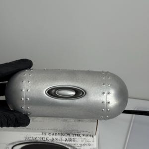 May include: A silver-colored, oval-shaped case with a riveted design and the Oakley logo. The case is resting on a box with the text "SCIENCE AND ART". The case is held by a gloved hand.