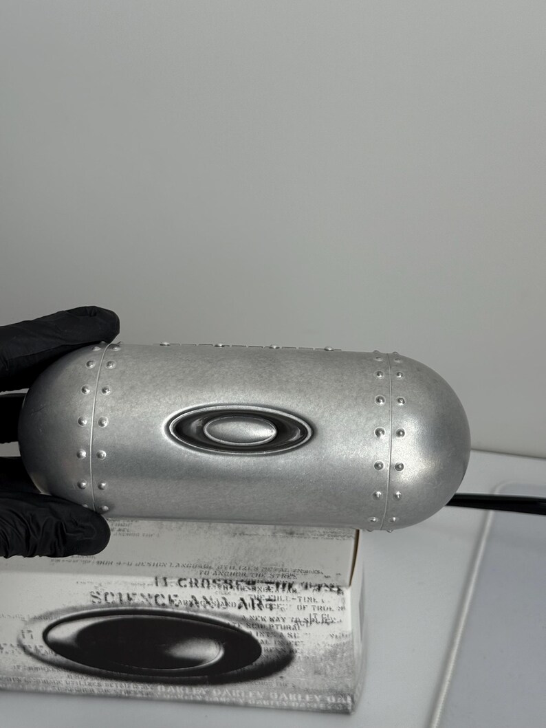 May include: A silver-colored, oval-shaped case with a riveted design and the Oakley logo. The case is resting on a box with the text "SCIENCE AND ART". The case is held by a gloved hand.