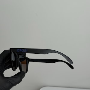 May include: Black Oakley sunglasses with dark lenses. The brand name is printed in blue on the left arm. The sunglasses are held by a gloved hand.