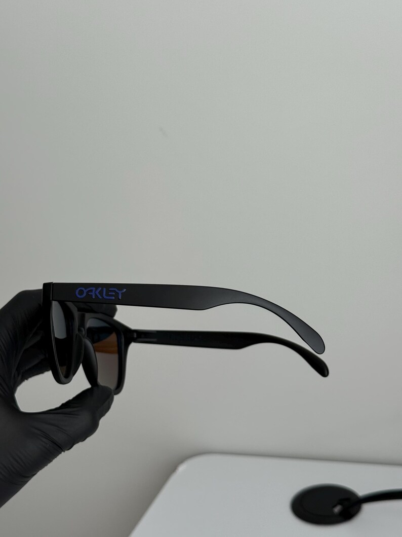 May include: Black Oakley sunglasses with dark lenses. The brand name is printed in blue on the left arm. The sunglasses are held by a gloved hand.