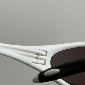 May include: Close-up of white sunglasses with a black arm and brown lenses. The sunglasses have a modern design with a distinctive hinge detail. The frame is white, and the lenses are a dark brown color.