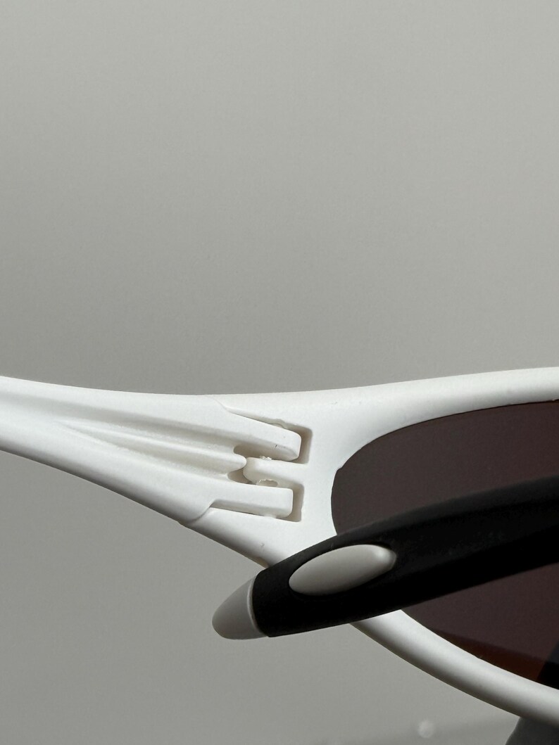 May include: Close-up of white sunglasses with a black arm and brown lenses. The sunglasses have a modern design with a distinctive hinge detail. The frame is white, and the lenses are a dark brown color.