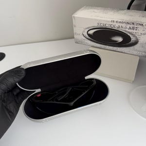 May include: Black sunglasses inside a silver case with a black interior. The case is open, revealing the sunglasses. A white box with the text "SCIENCE AND ART" is in the background.