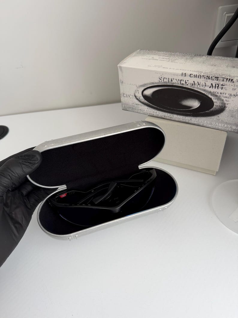 May include: Black sunglasses inside a silver case with a black interior. The case is open, revealing the sunglasses. A white box with the text "SCIENCE AND ART" is in the background.
