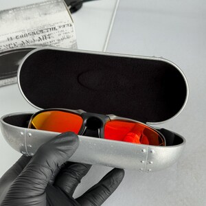 May include: Black sunglasses with red lenses rest in an open, silver-colored case. The case has a black interior and is held by a gloved hand. A box with text is visible in the background.