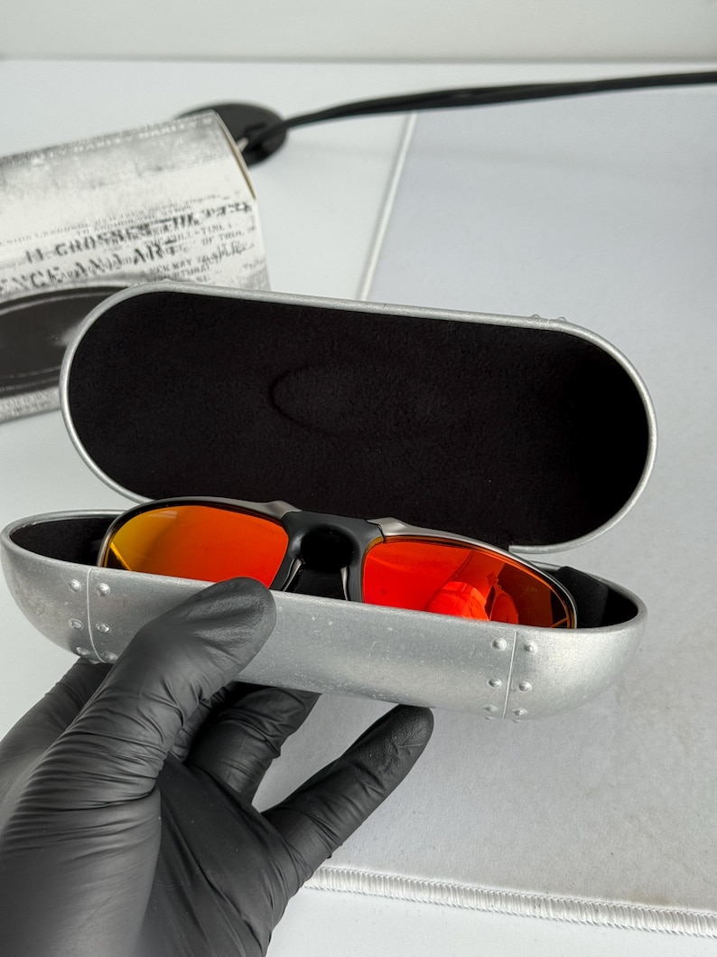 May include: Black sunglasses with red lenses rest in an open, silver-colored case. The case has a black interior and is held by a gloved hand. A box with text is visible in the background.