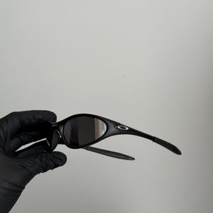K&ouml;nnte beinhalten: Schwarze Sonnenbrille mit geschwungenem Rahmen und dunklen Gl&auml;sern. Die Sonnenbrille hat ein schlankes Design mit einem kleinen Logo am B&uuml;gel. Die B&uuml;gel sind d&uuml;nn und reichen bis zu den Ohren.