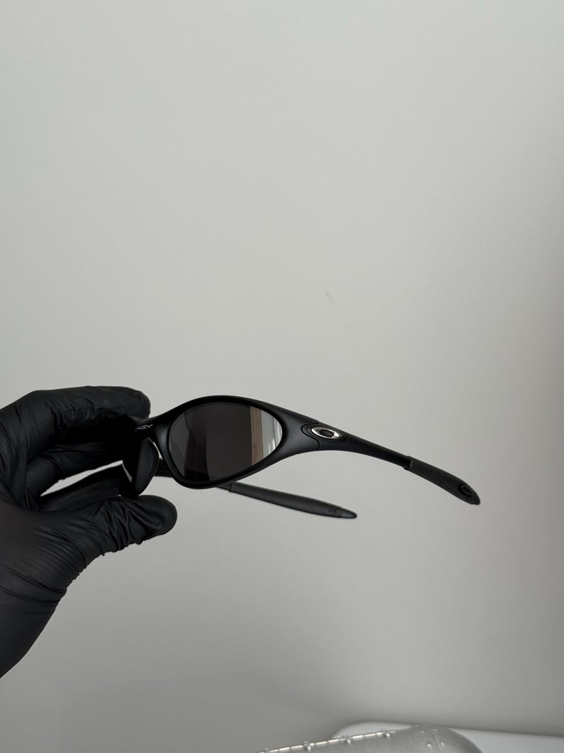 K&ouml;nnte beinhalten: Schwarze Sonnenbrille mit geschwungenem Rahmen und dunklen Gl&auml;sern. Die Sonnenbrille hat ein schlankes Design mit einem kleinen Logo am B&uuml;gel. Die B&uuml;gel sind d&uuml;nn und reichen bis zu den Ohren.