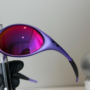 May include: Close-up of purple sunglasses with red-tinted lenses. The sunglasses have a sleek, curved design with black accents on the arms. The lenses reflect a vibrant pink hue, and the text "PRIZM P" is visible on the lens.