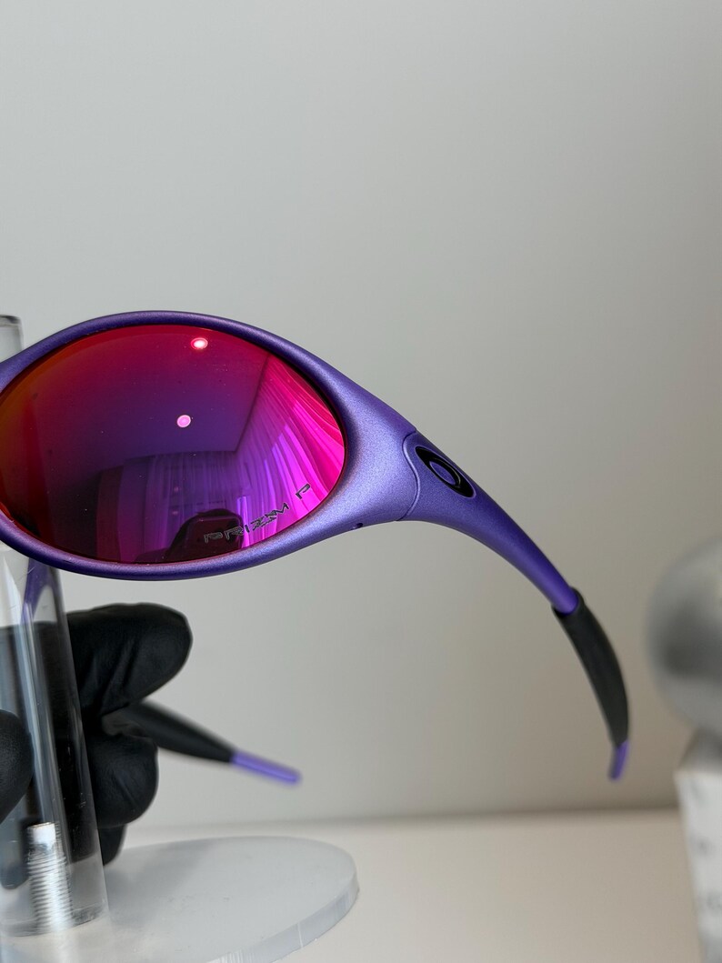 May include: Close-up of purple sunglasses with red-tinted lenses. The sunglasses have a sleek, curved design with black accents on the arms. The lenses reflect a vibrant pink hue, and the text "PRIZM P" is visible on the lens.