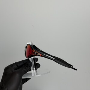 May include: Black sunglasses with red lenses and flame-like orange and black accents on the temples. The sunglasses are displayed on a clear stand against a white background.