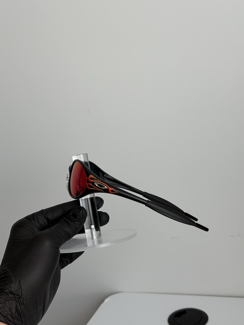 May include: Black sunglasses with red lenses and flame-like orange and black accents on the temples. The sunglasses are displayed on a clear stand against a white background.