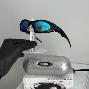 May include: Black and blue Oakley sunglasses with reflective blue lenses. The sunglasses are displayed on a clear stand, with a silver case and a white box with text visible below.