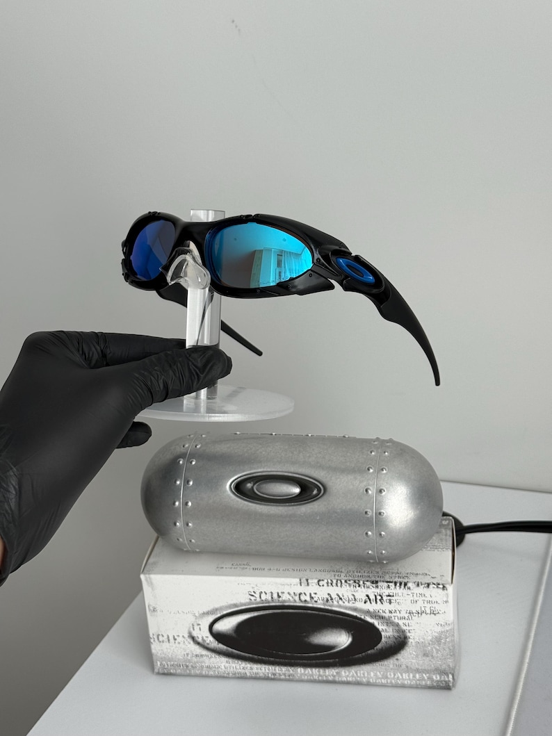 May include: Black and blue Oakley sunglasses with reflective blue lenses. The sunglasses are displayed on a clear stand, with a silver case and a white box with text visible below.