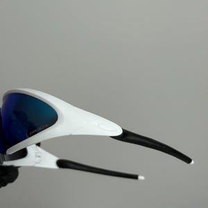 Puede incluir: Gafas de sol deportivas blancas y negras con lentes tintadas en azul. Las gafas tienen un dise&ntilde;o elegante con la palabra "PRIZM" impresa en la lente. Las patillas son negras con puntas blancas.