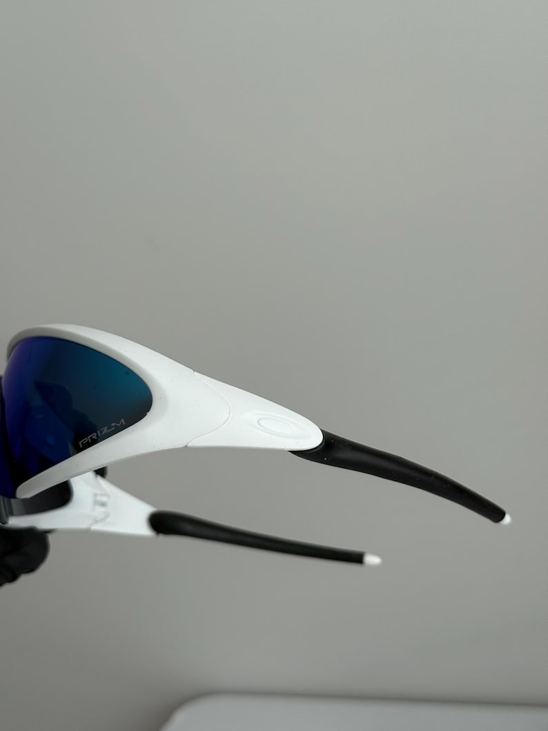 Puede incluir: Gafas de sol deportivas blancas y negras con lentes tintadas en azul. Las gafas tienen un dise&ntilde;o elegante con la palabra "PRIZM" impresa en la lente. Las patillas son negras con puntas blancas.
