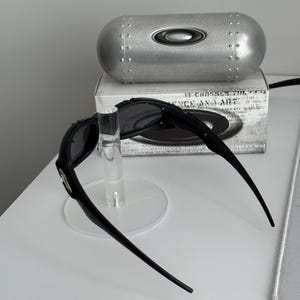 May include: Black sunglasses with a silver case and a clear display stand. The sunglasses have a sleek, modern design with dark lenses and a curved frame. The silver case has the brand logo on it.