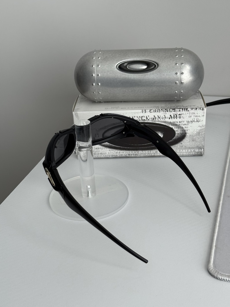 May include: Black sunglasses with a silver case and a clear display stand. The sunglasses have a sleek, modern design with dark lenses and a curved frame. The silver case has the brand logo on it.