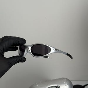 Puede incluir: Gafas de sol plateadas con lentes oscuras. Las gafas tienen un dise&ntilde;o elegante con patillas curvas y una forma de lente peque&ntilde;a y redondeada. Tambi&eacute;n se ve un estuche plateado con el logotipo de la marca.