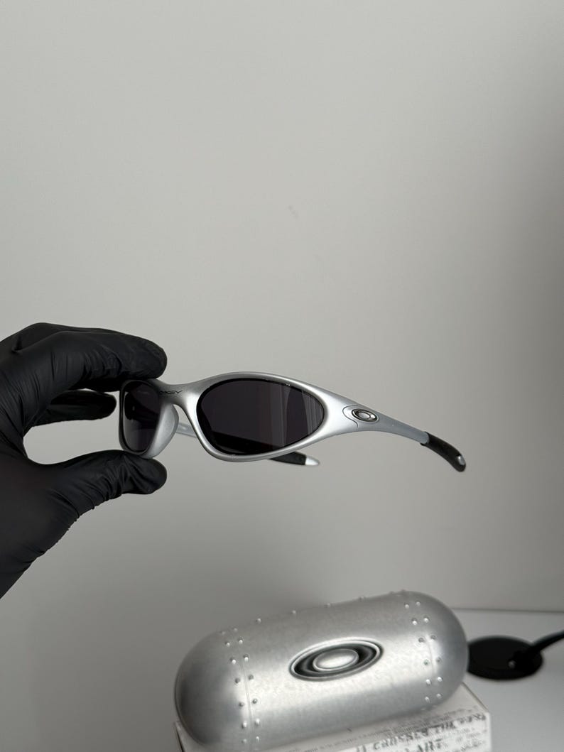 Puede incluir: Gafas de sol plateadas con lentes oscuras. Las gafas tienen un dise&ntilde;o elegante con patillas curvas y una forma de lente peque&ntilde;a y redondeada. Tambi&eacute;n se ve un estuche plateado con el logotipo de la marca.