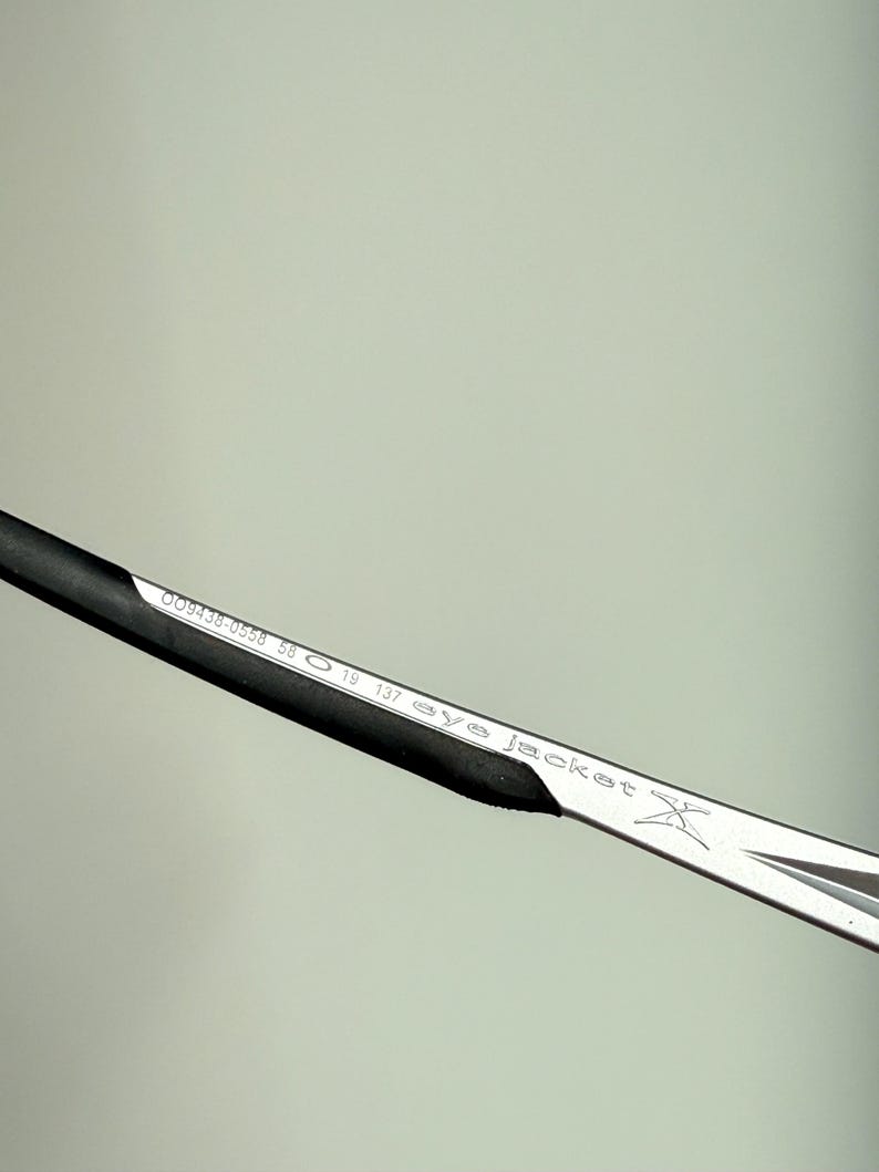May include: Close-up of a silver and black eyeglass frame. The frame has the text "eye jacket" and other numbers and letters printed on it. The frame is angled diagonally across the image.