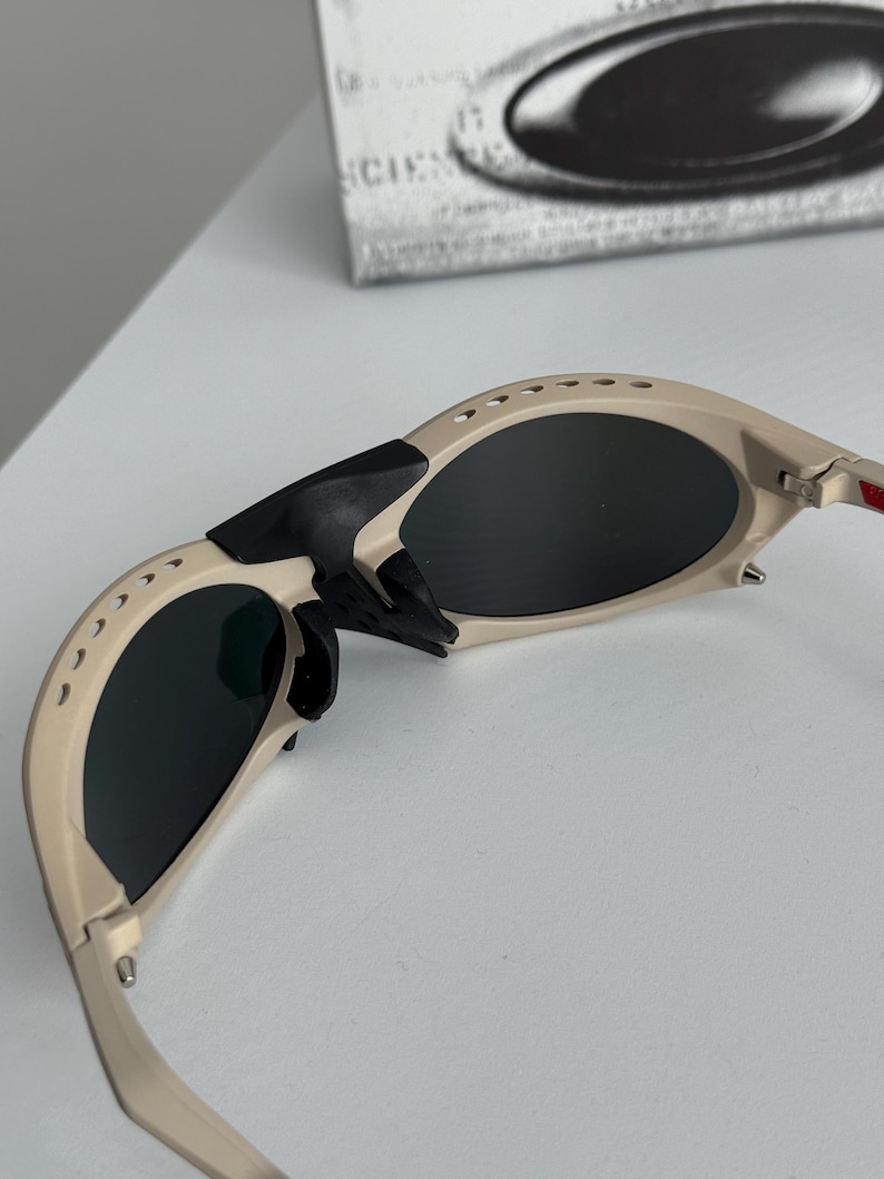 Może przedstawiać: Beżowe okulary przeciwsłoneczne z ciemnymi soczewkami. Okulary mają sportowy design z zakrzywioną oprawką i czarnym noskiem. Soczewki są ciemne, a oprawka ma małe otwory wentylacyjne. Okulary są ustawione pod kątem, pokazując szczeg&oacute;ły oprawki i soczewek.