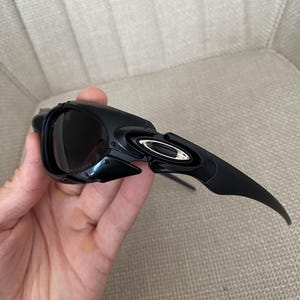 May include: Black sunglasses with a sleek, modern design. The sunglasses feature dark lenses and a curved frame with a metallic accent. The arms of the sunglasses are long and slightly curved.